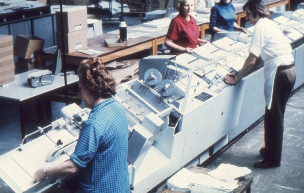 Man sieht auf einem Bild aus dem Jahr 1975 eine Zeile Sortier- und Verpackungsmaschinen, an denen Lagerarbeiter gerade Bücher und andere Studienmaterialien für den Versand vorbereiten.